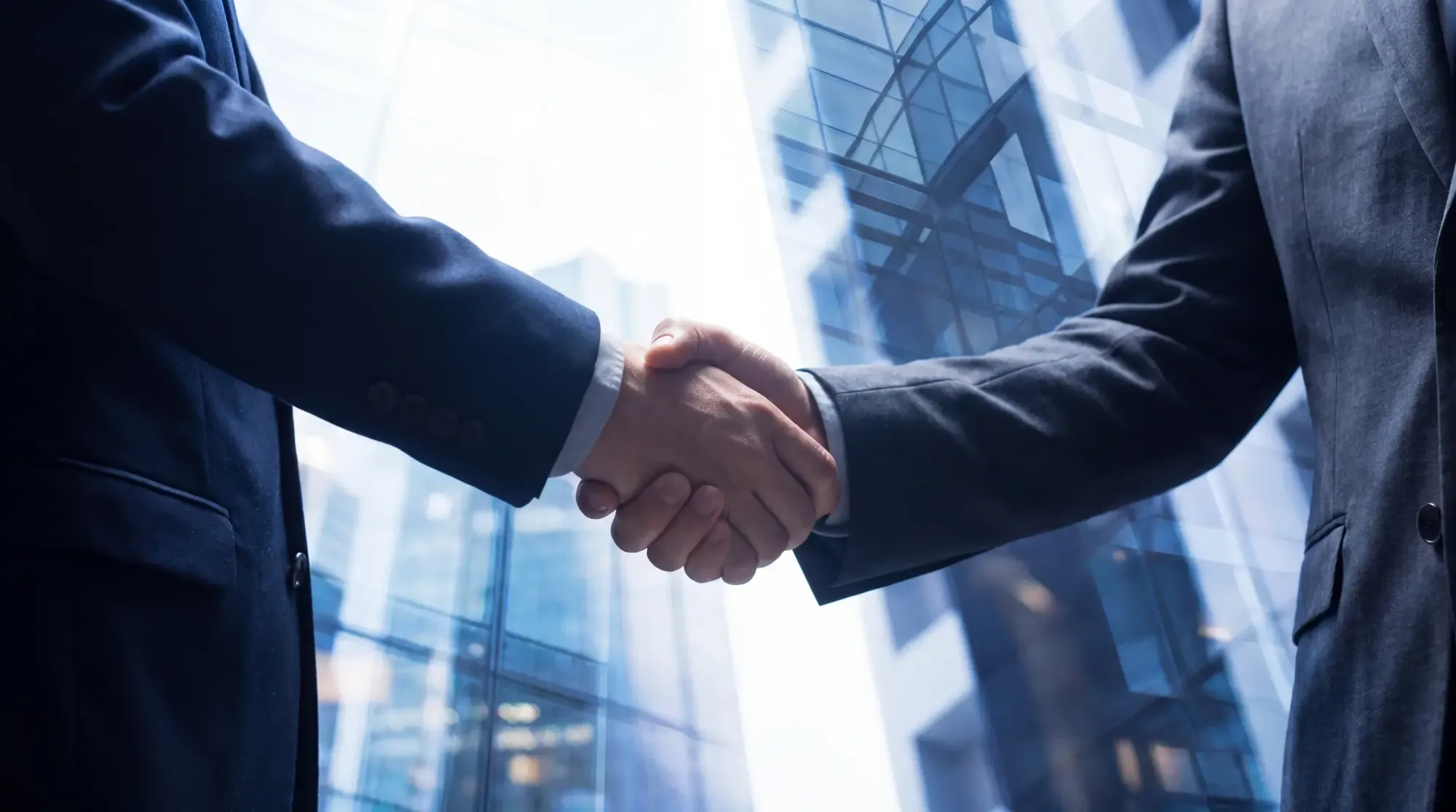 Two businessmen in suits shaking hands, symbolizing agreement with blurred corporate building